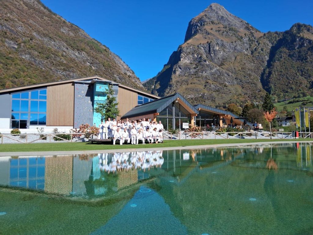 Gruppo in gi posa davanti al centro sportivo di Olivone, con laghetto e montagne sullo sfondo